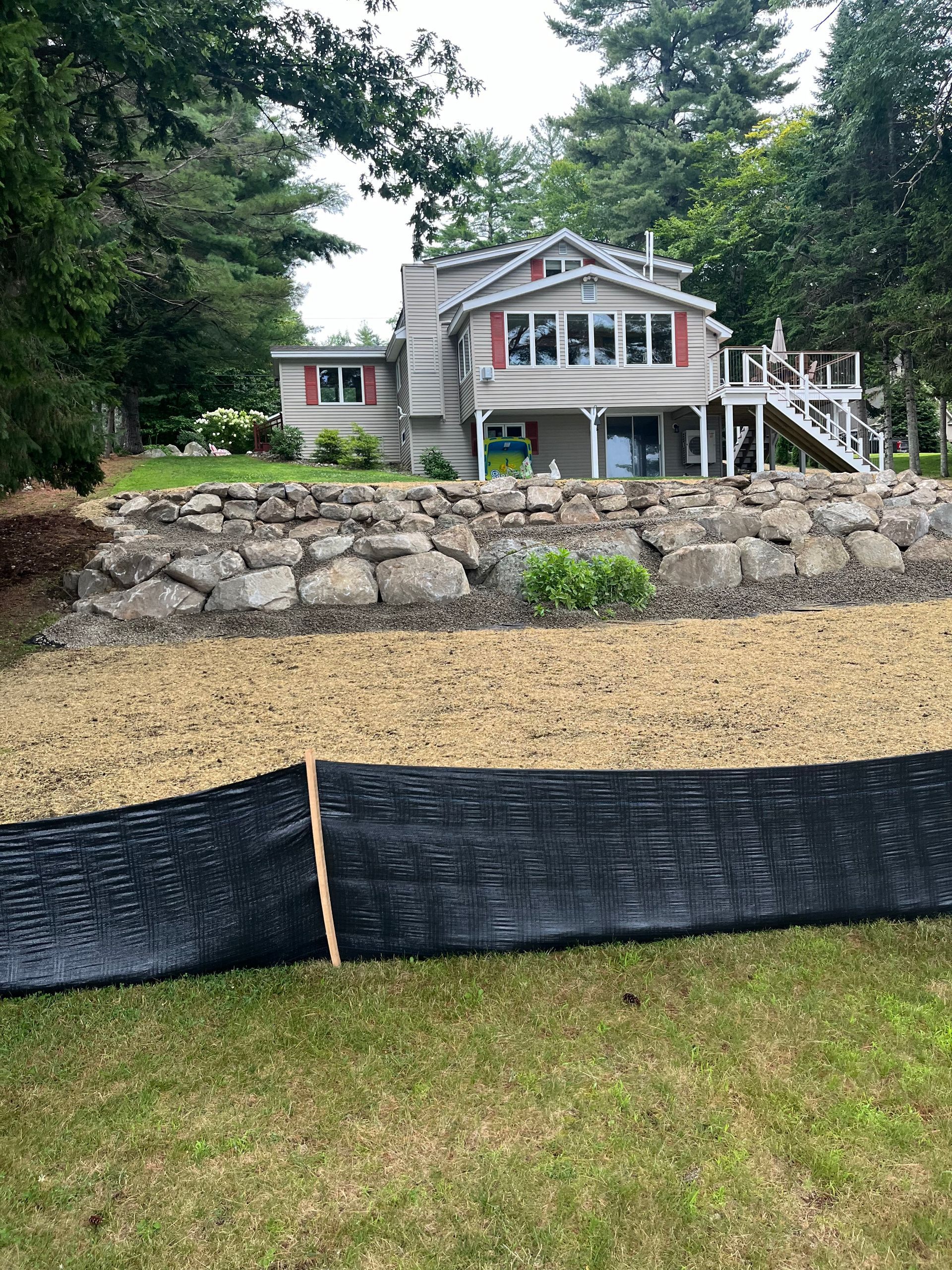 A light-colored house with red shutters sits behind a stone retaining wall, mulch, and a black silt fence in a yard.