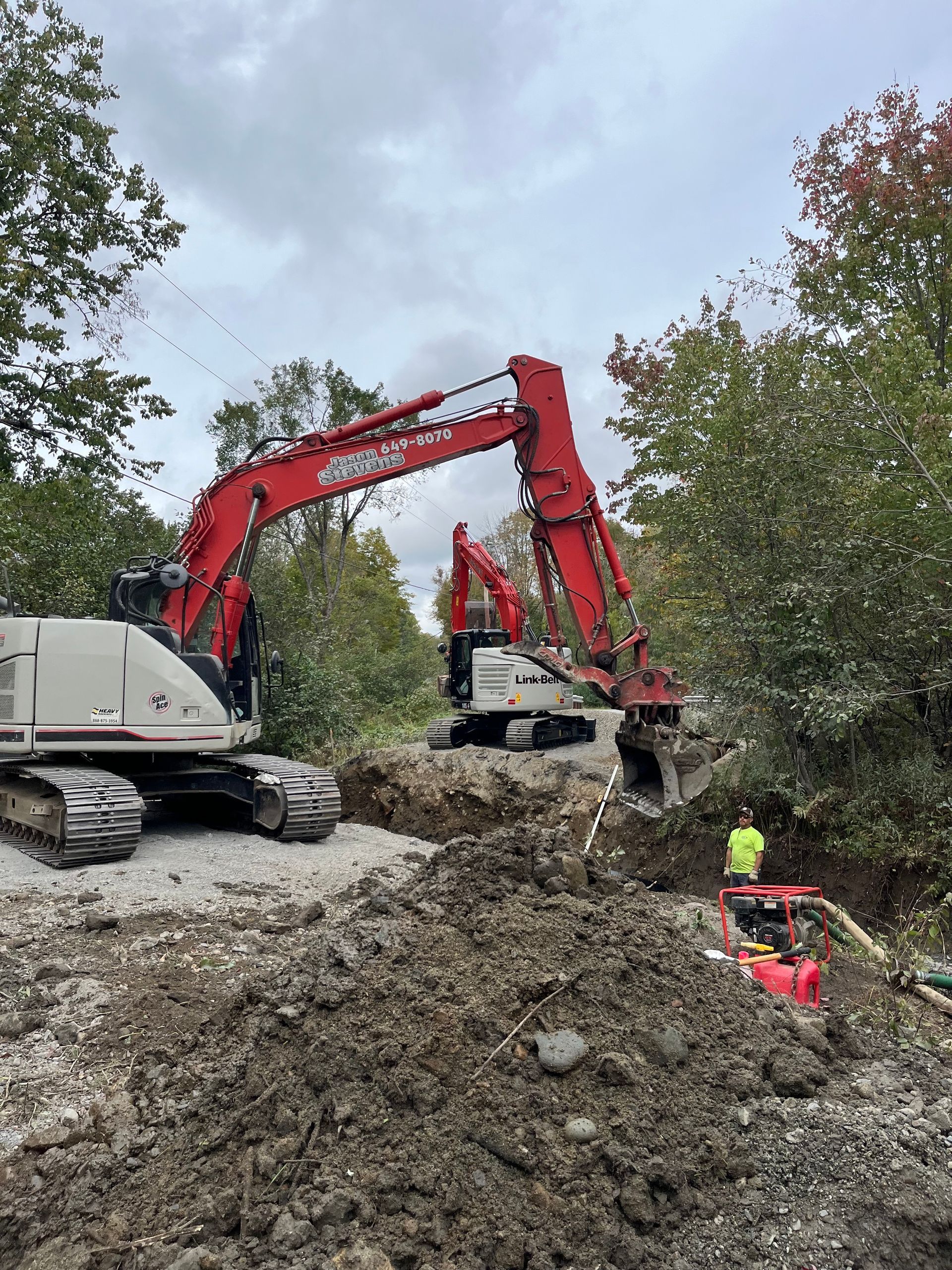Two red excavators work on a construction site with piles of dirt and a worker standing nearby.