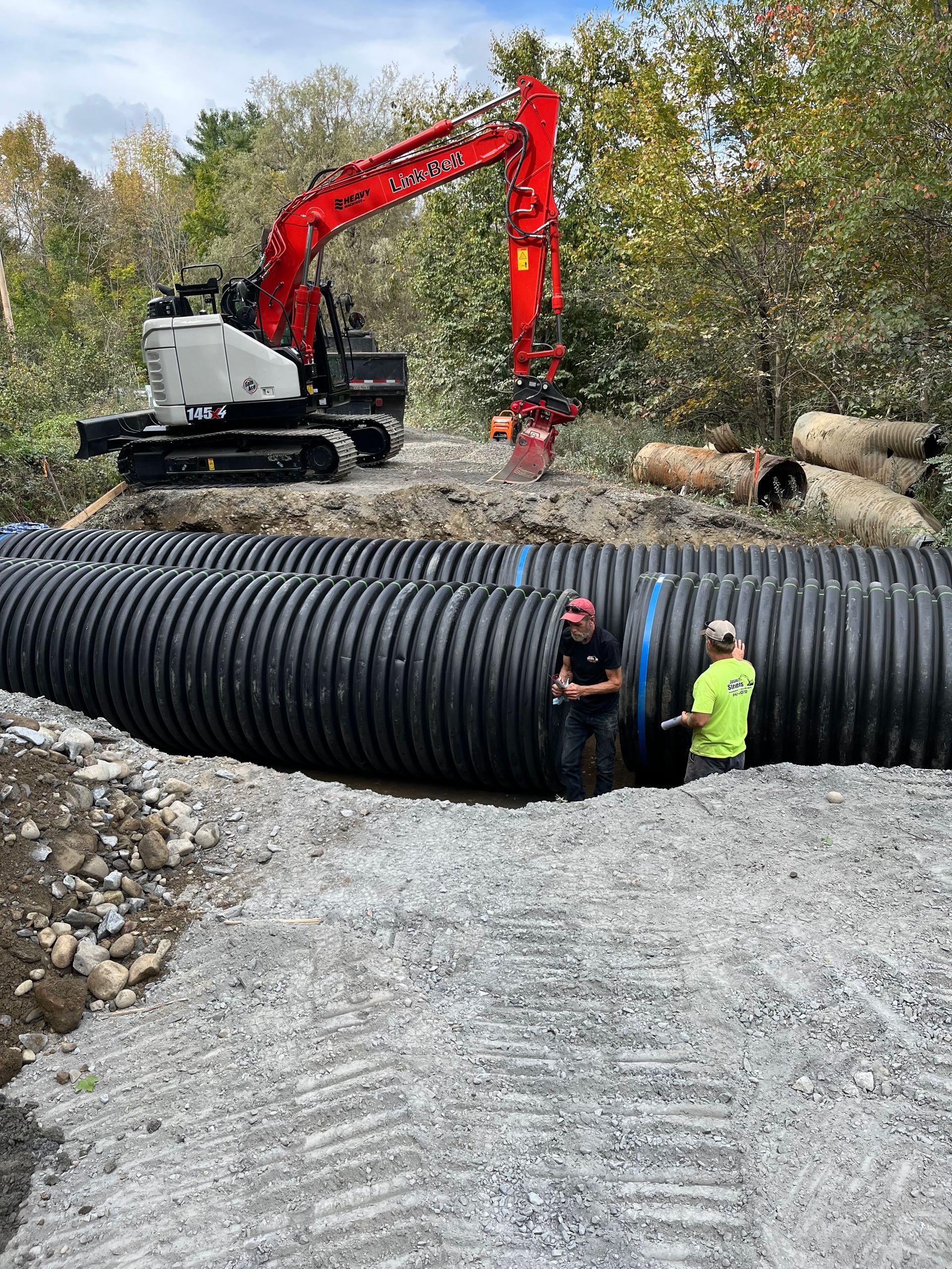 Two workers stand by large, black corrugated pipes at a construction site with a red excavator in the background.