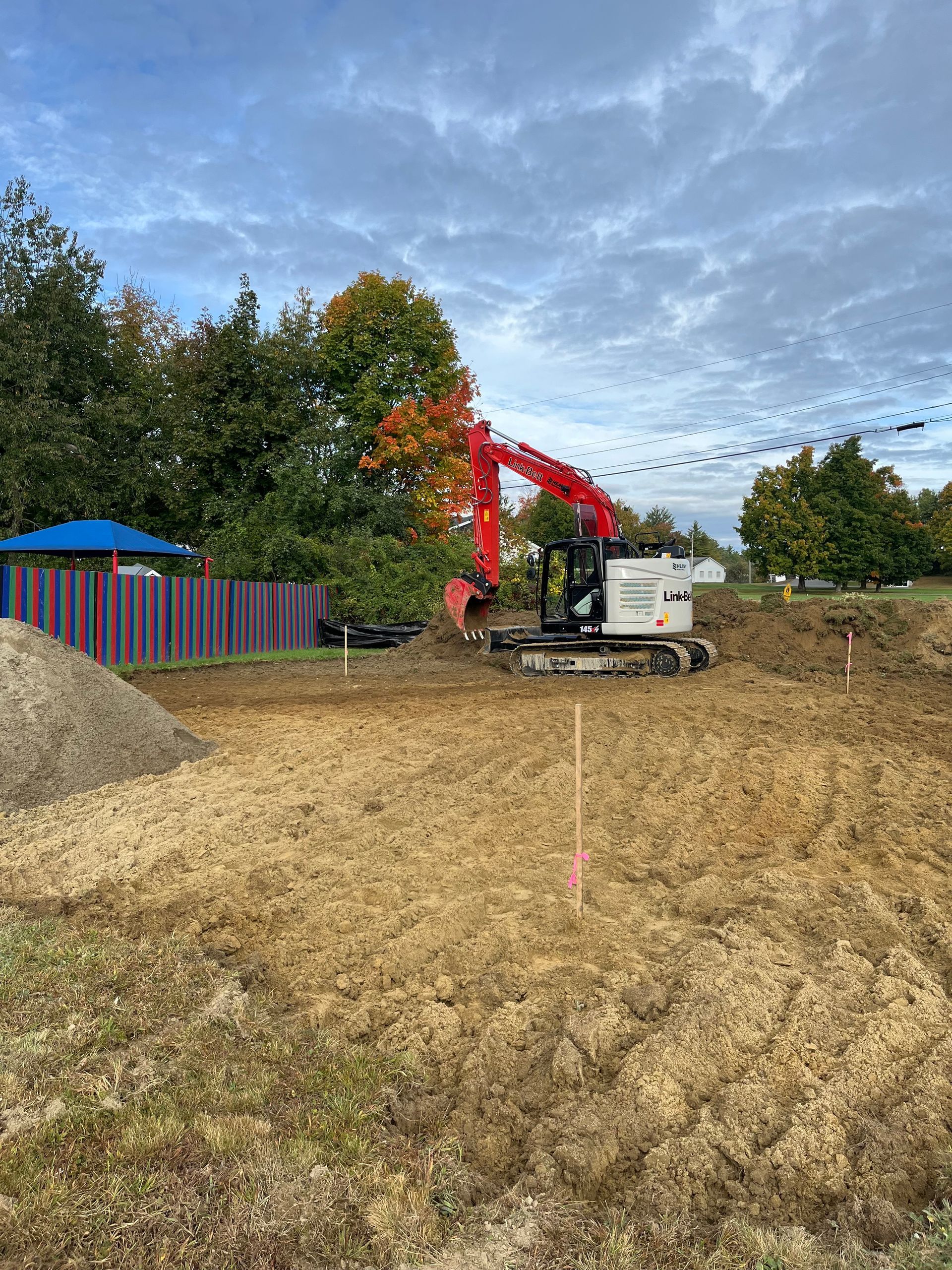 A red excavator works on an excavation site with a pile of gravel and a colorful fence nearby under a cloudy sky.