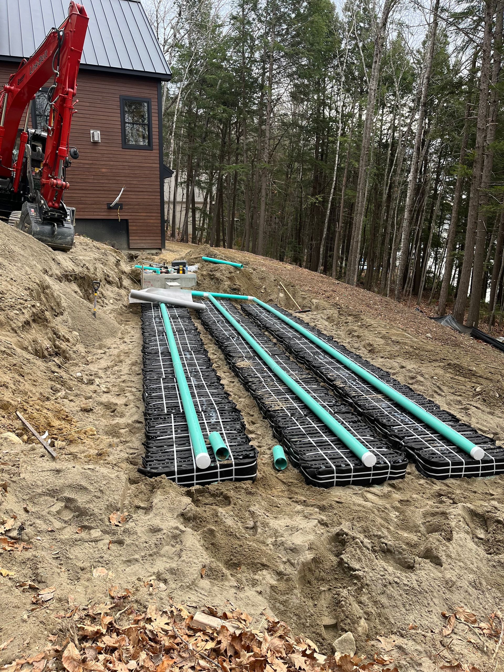 Construction of a septic system leach field with green pipes laid over black drainage chambers near a house and woods.