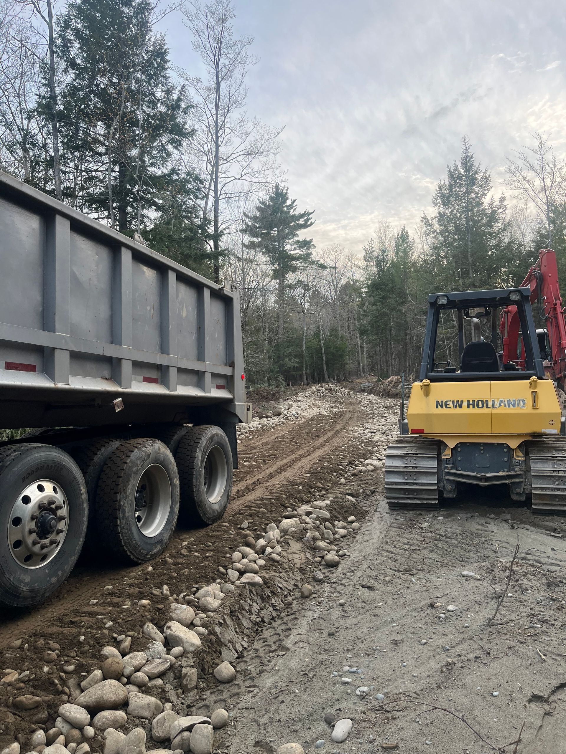 A dump truck and a yellow bulldozer parked on a dirt road in a wooded area.
