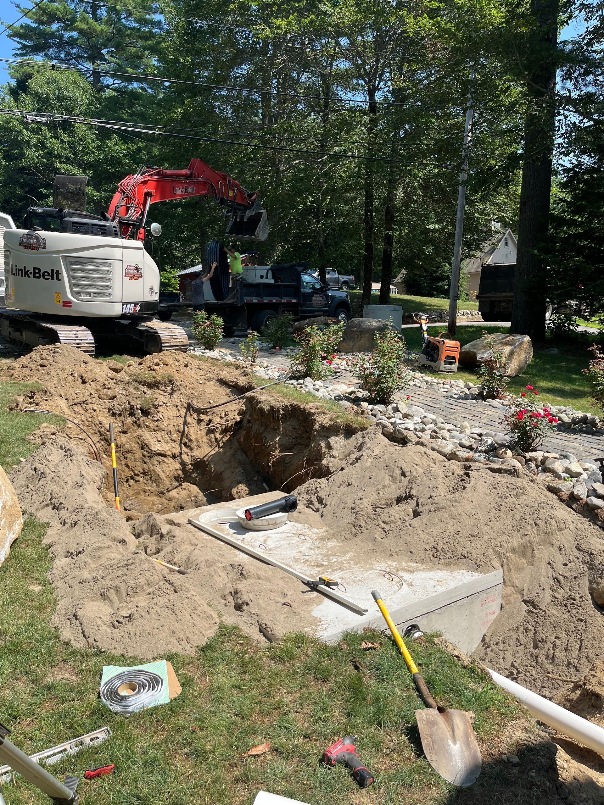 A red excavator digs a trench in a grassy yard, exposing a concrete structure with tools placed on the ground nearby.