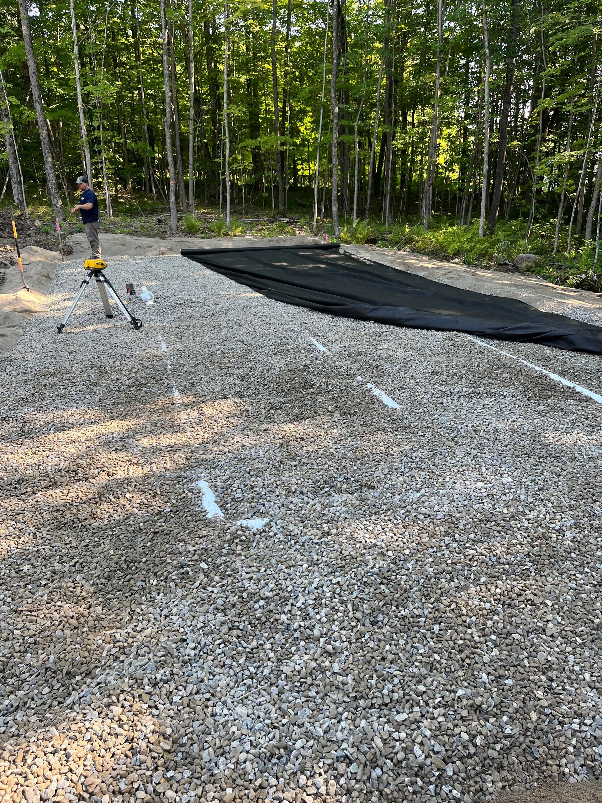 A person uses a surveying tool on a gravel construction site with a section of black landscape fabric laid out.