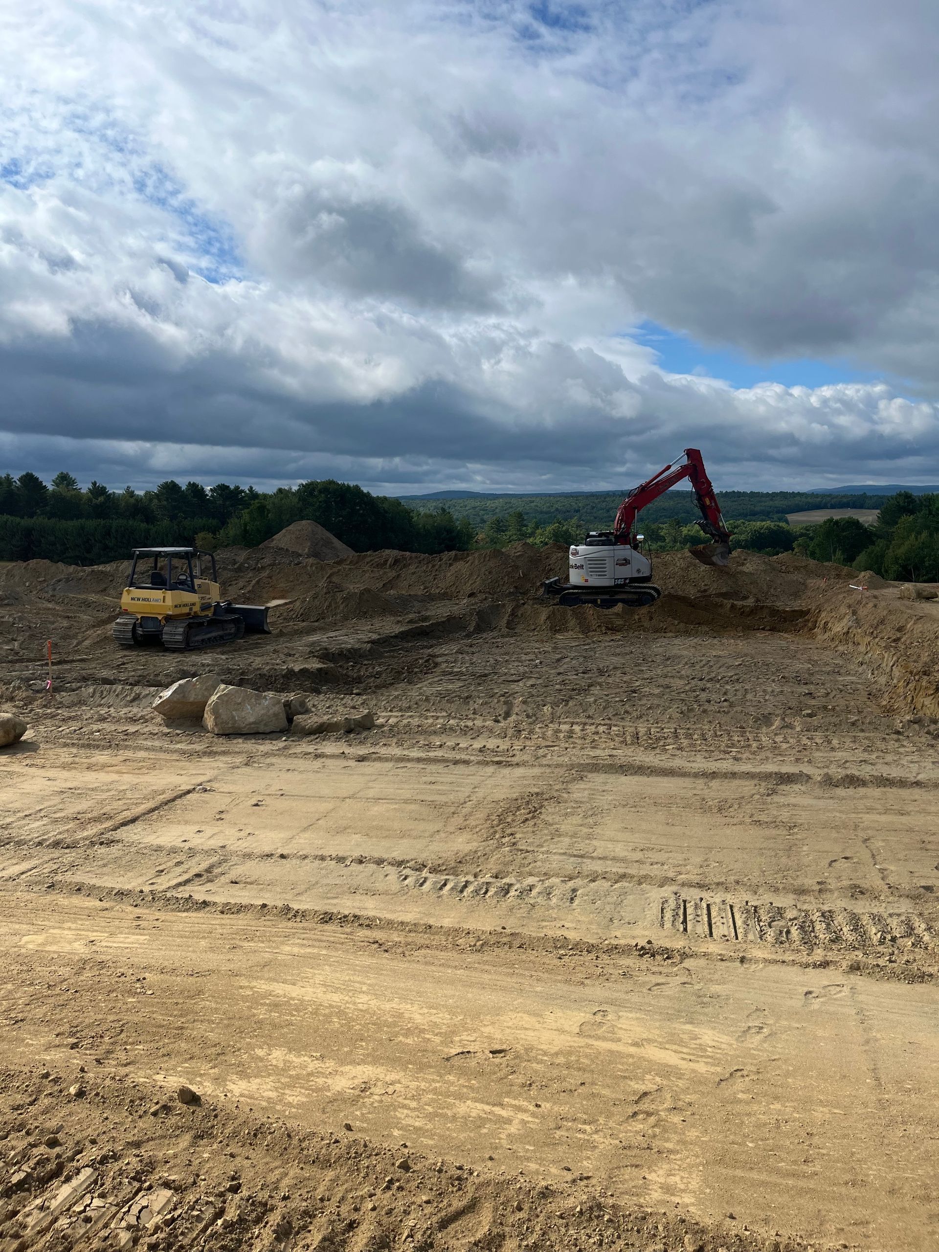 A construction site with two excavators working on a cleared, earthy plot under a cloudy, expansive sky.