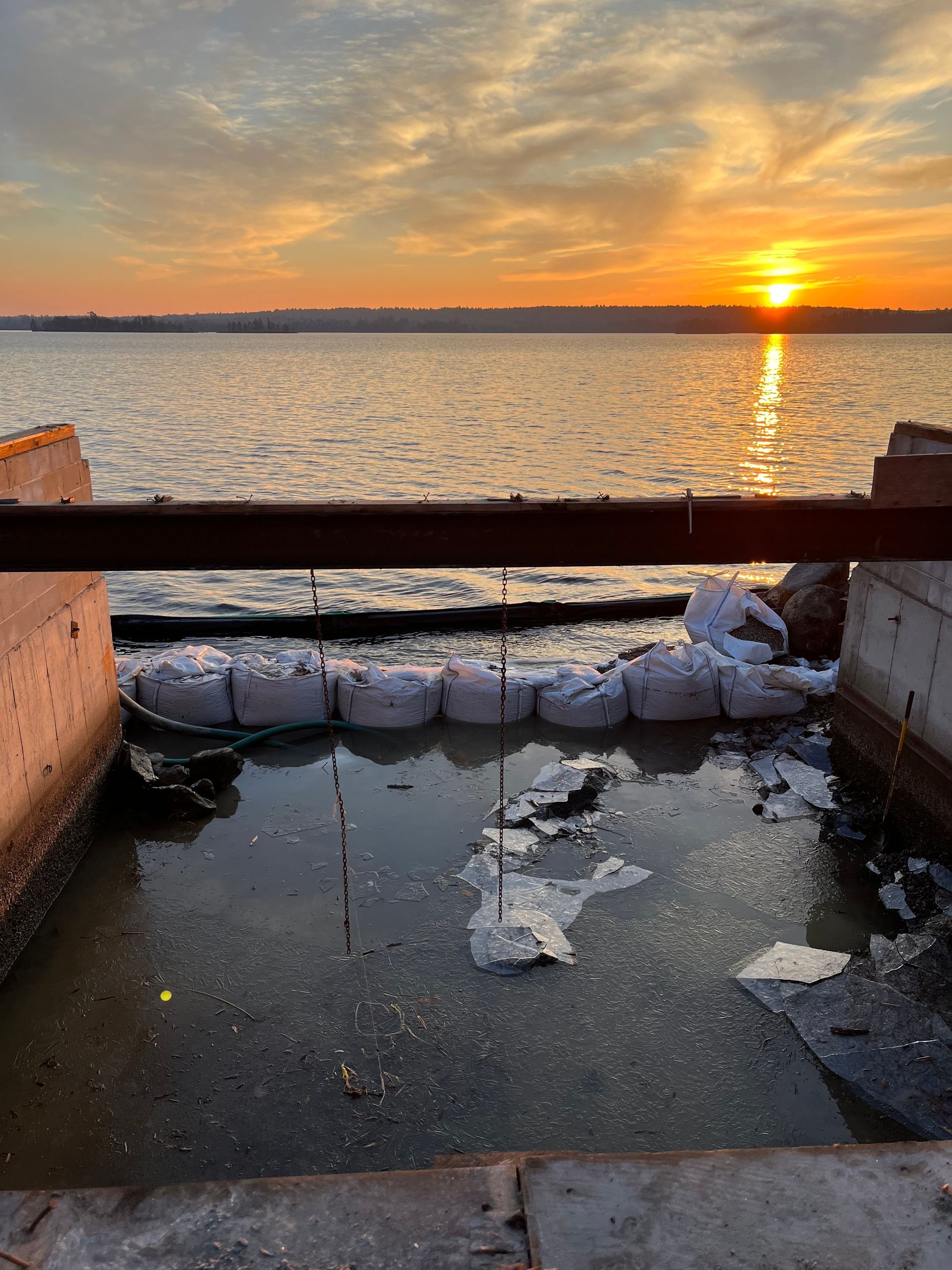 A sunset over a body of water, viewed past a metal rail and a line of white sandbags placed in a muddy area.