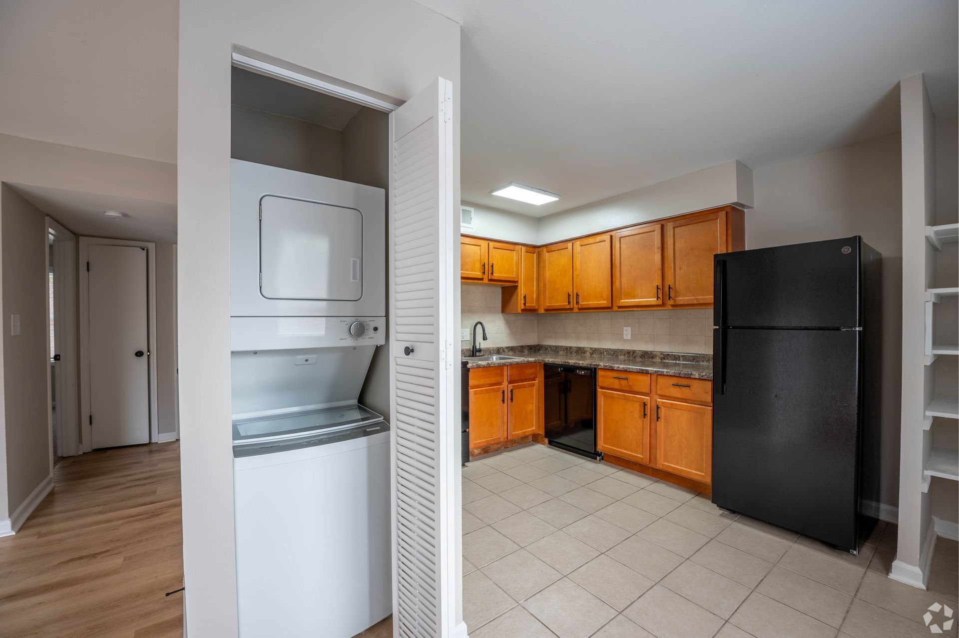 Interior view of a kitchen with washer/dryer unit, wooden cabinets, black refrigerator, and tiled floor.