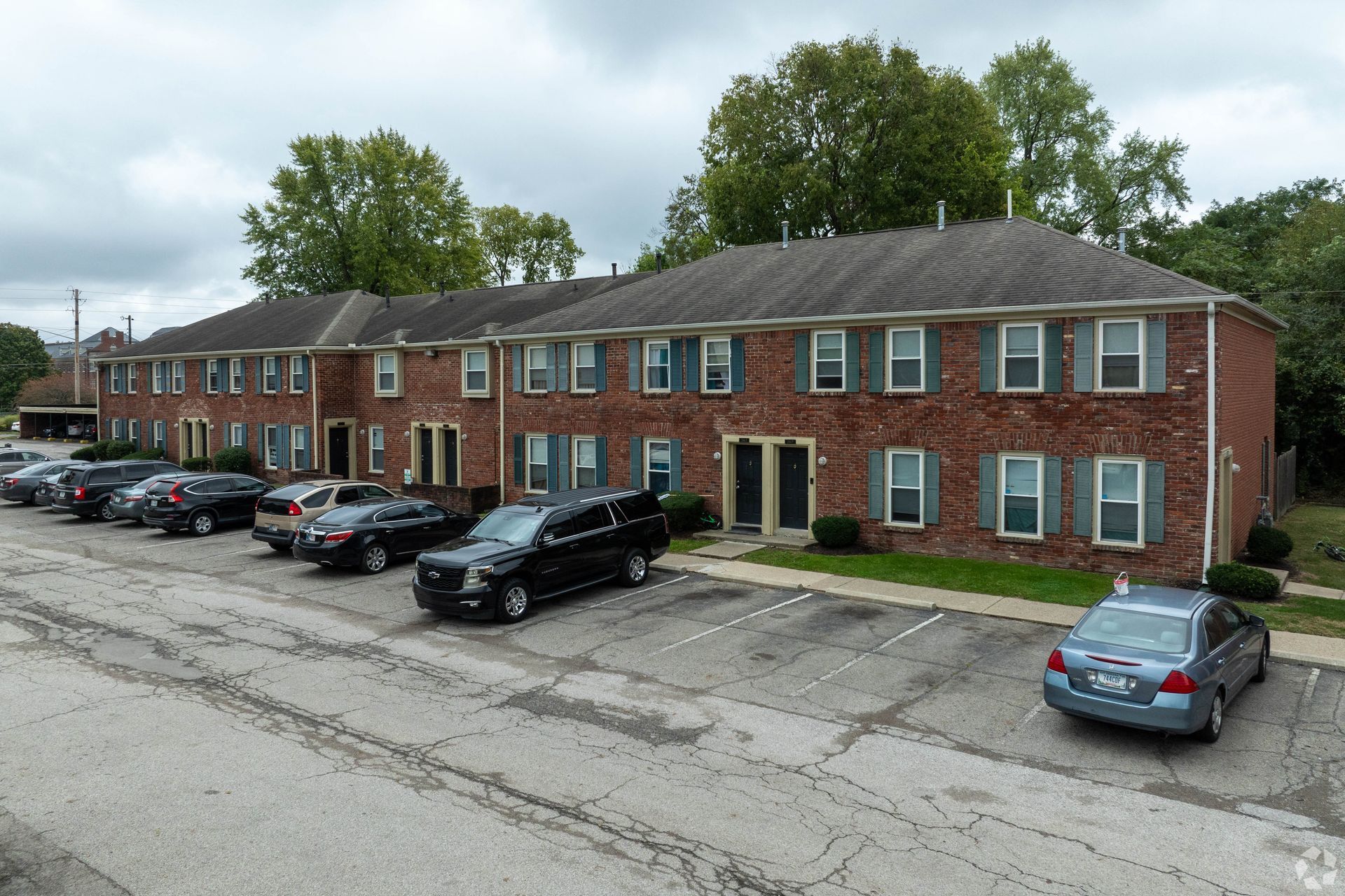 Brick apartment building with parked cars in a parking lot on a cloudy day.