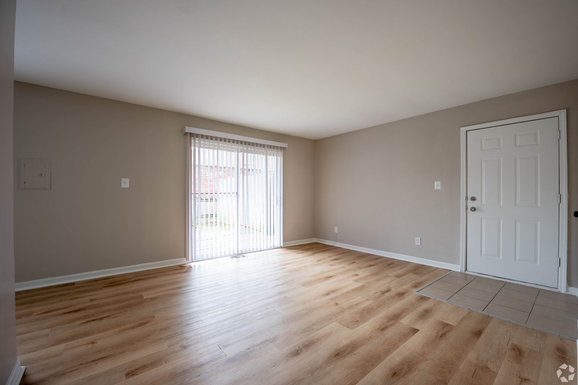 Empty living room with wood-look floors, sliding glass door with vertical blinds, and white entry door.