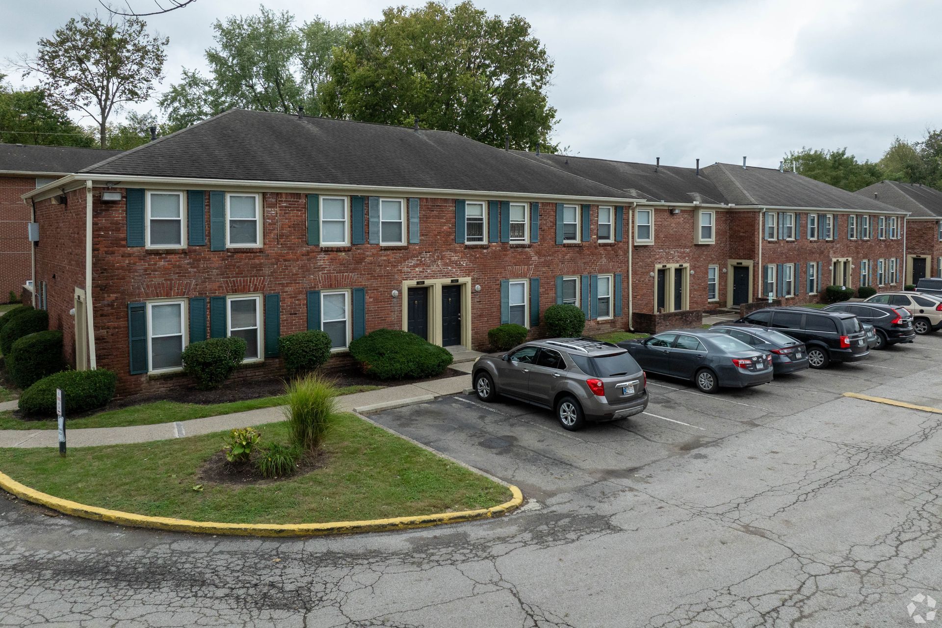 Red brick two-story apartment buildings with green shutters and parking in front, set against a cloudy sky.