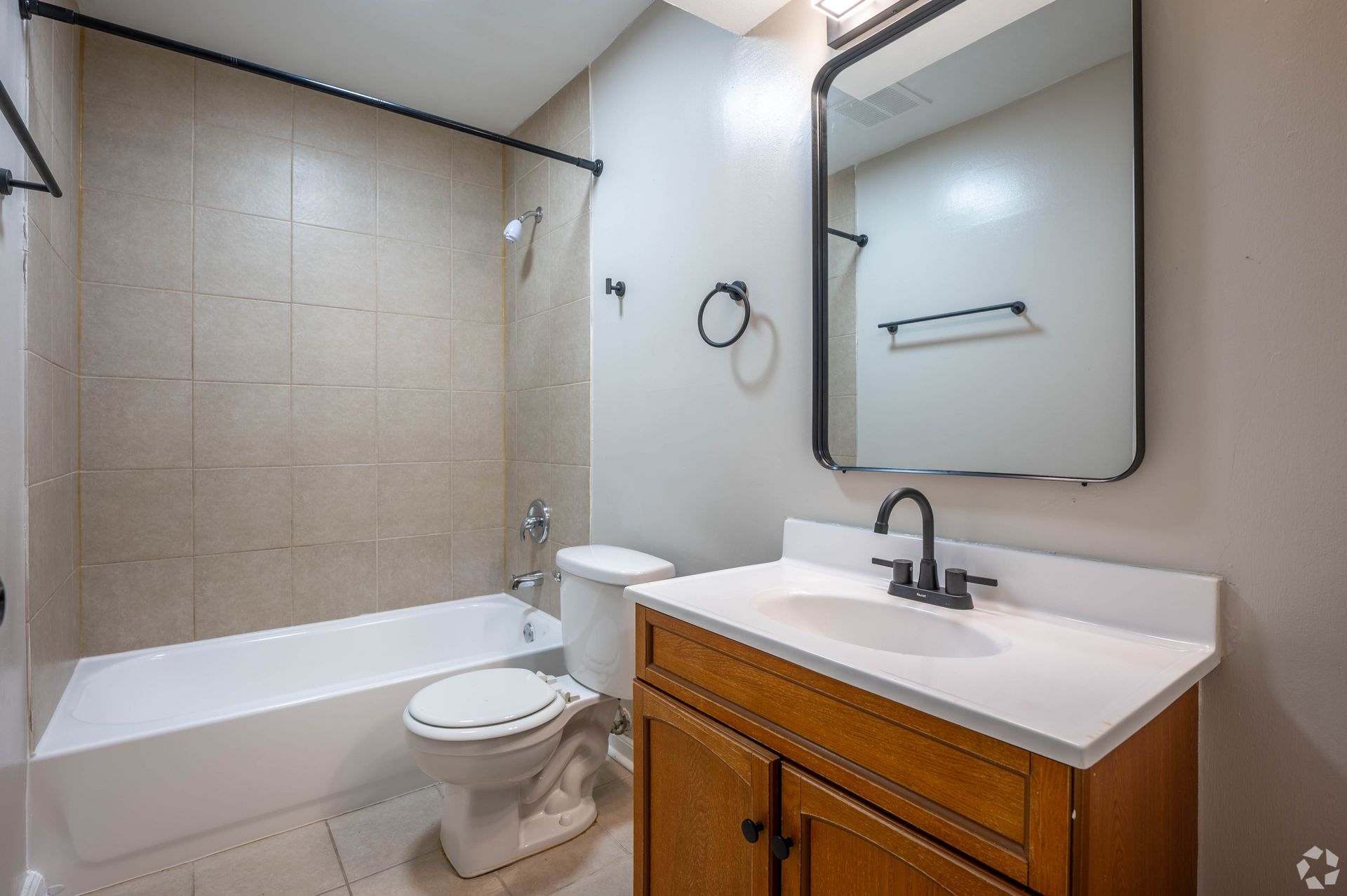 Bathroom with tan tile, white tub and toilet, wooden vanity, and black fixtures.