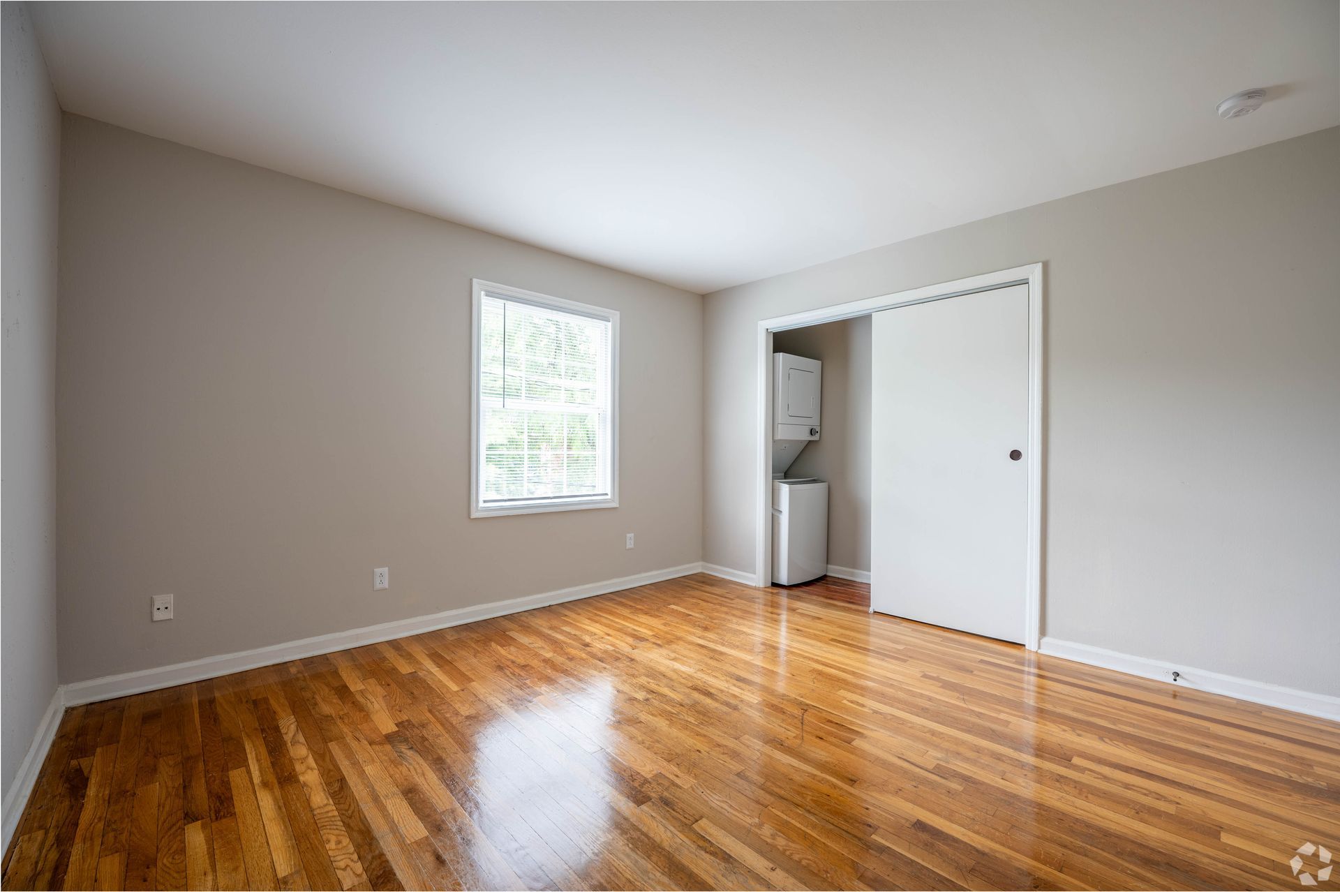 Empty bedroom with hardwood floors, a window, and a closet containing a stacked washer and dryer.