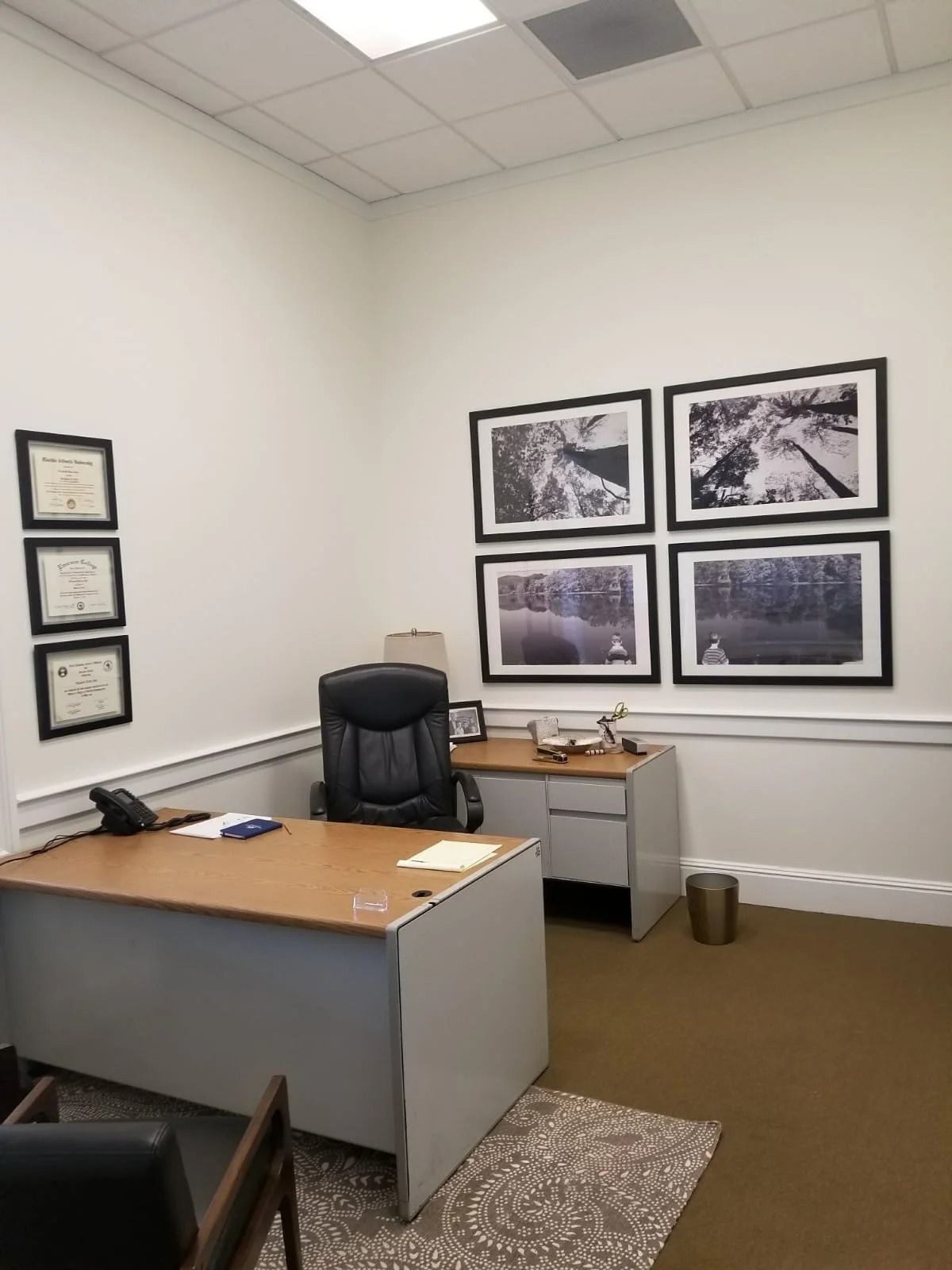 An office with a desk, chair, diplomas, and framed photos on the wall; a phone sits on the desk.
