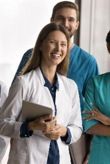 Healthcare workers smiling in a clinic hallway, one in a white coat holding a notebook.