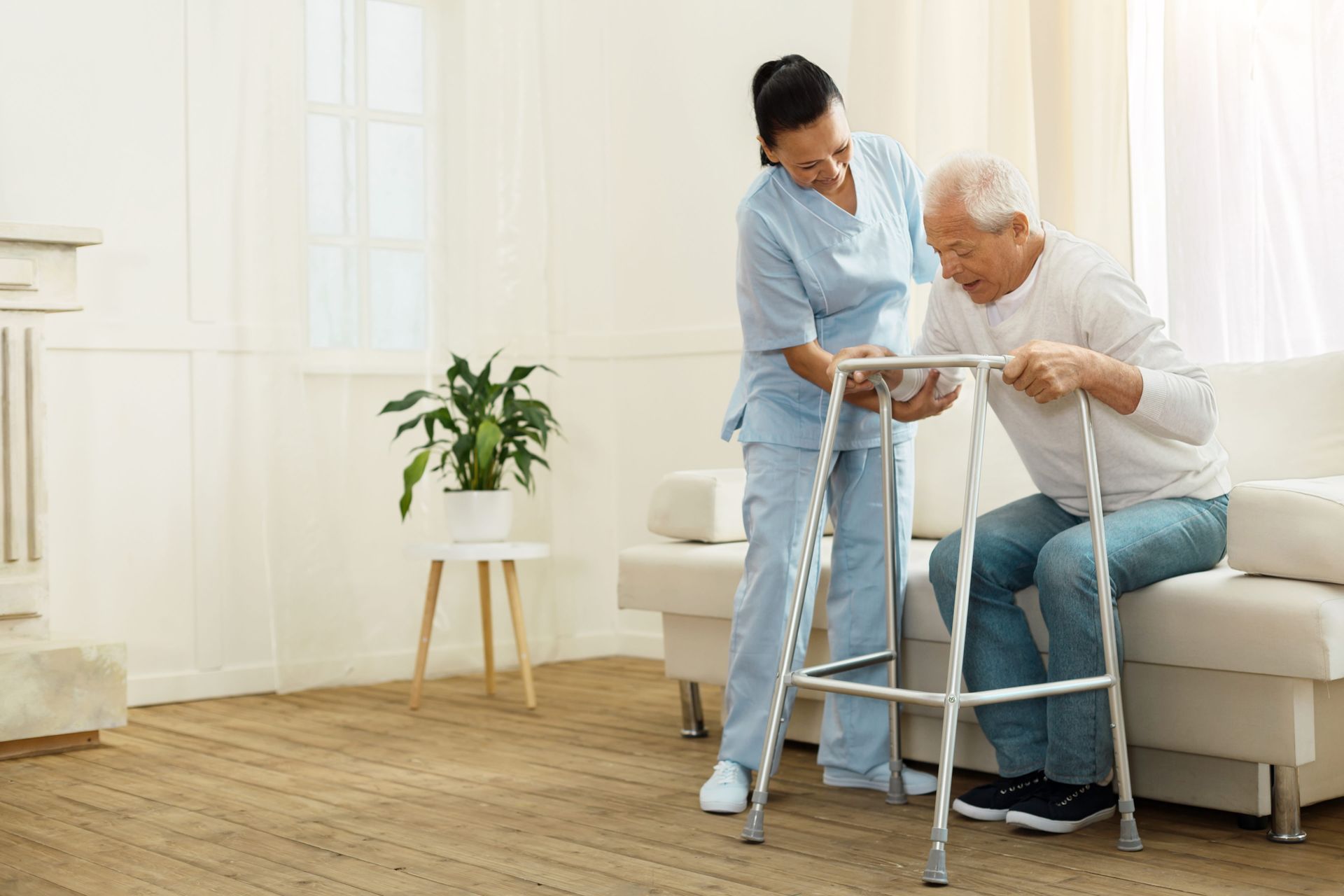 Caretaker helping an elderly person walk with a walker in a bright living room
