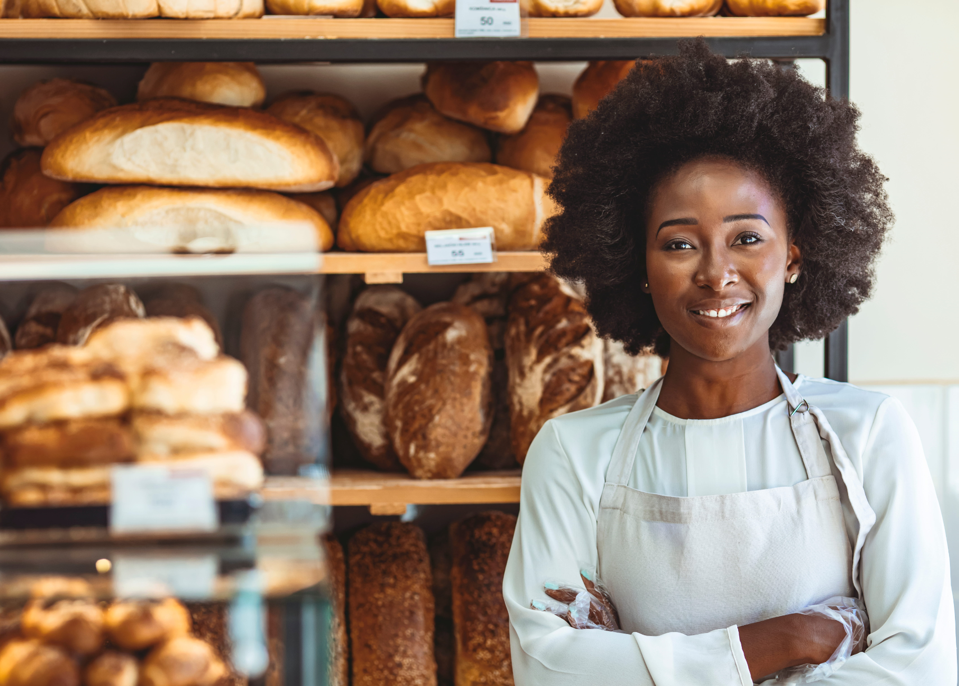Bakery worker in a white apron standing with arms crossed in front of bread shelves