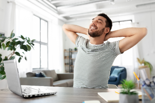 Man stretching at a desk beside a laptop in a bright home office.