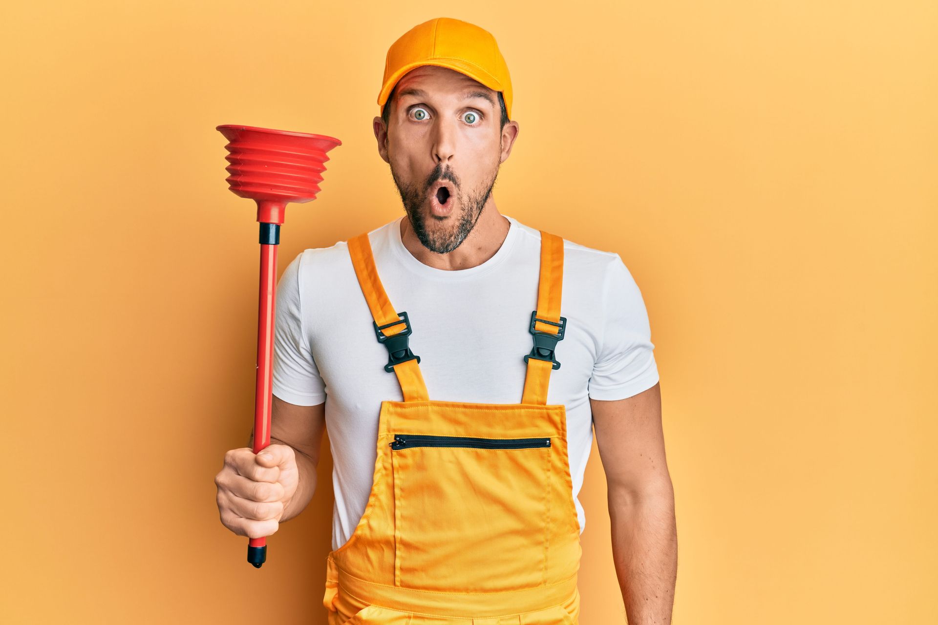 Plumber in orange overalls and cap holding a red plunger, looking surprised against a yellow background.