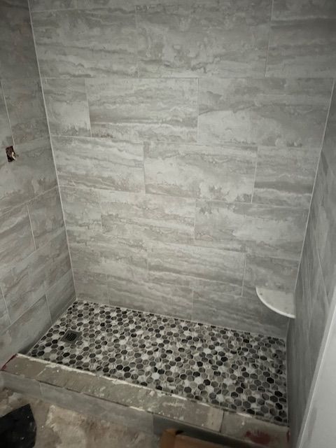 Shower stall with gray tile walls and pebble floor. A built-in shelf is in the corner.