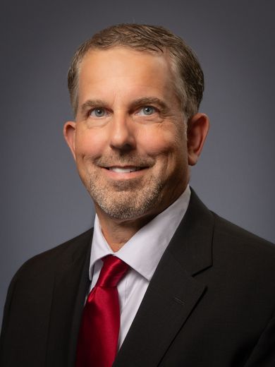 Headshot of a man with short brown hair and a beard, wearing a black suit, white shirt, and red necktie.