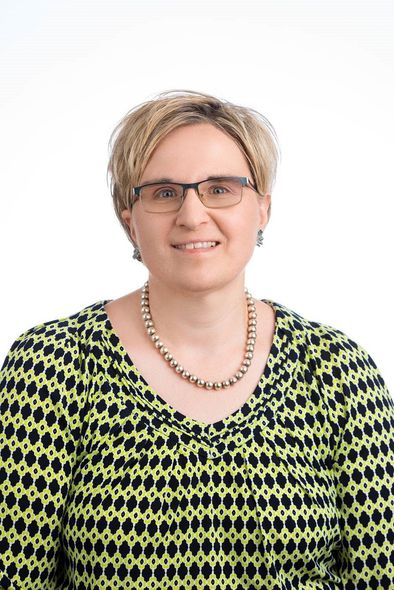 A person with short, light-brown hair wearing glasses and a lime-green and black patterned shirt with a beaded necklace.