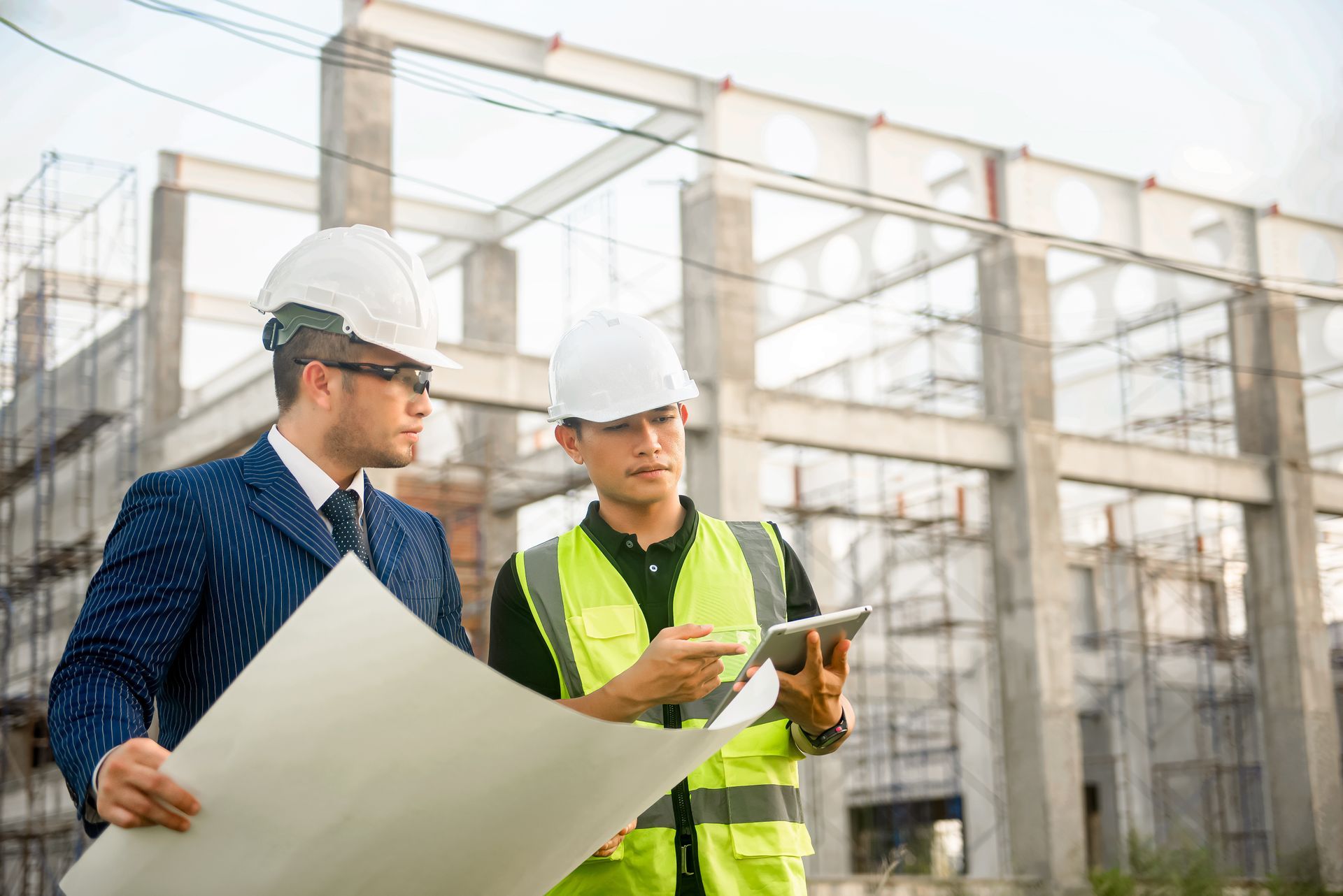 Two professionals in hard hats review building plans on paper and a tablet at a construction site with steel framework.