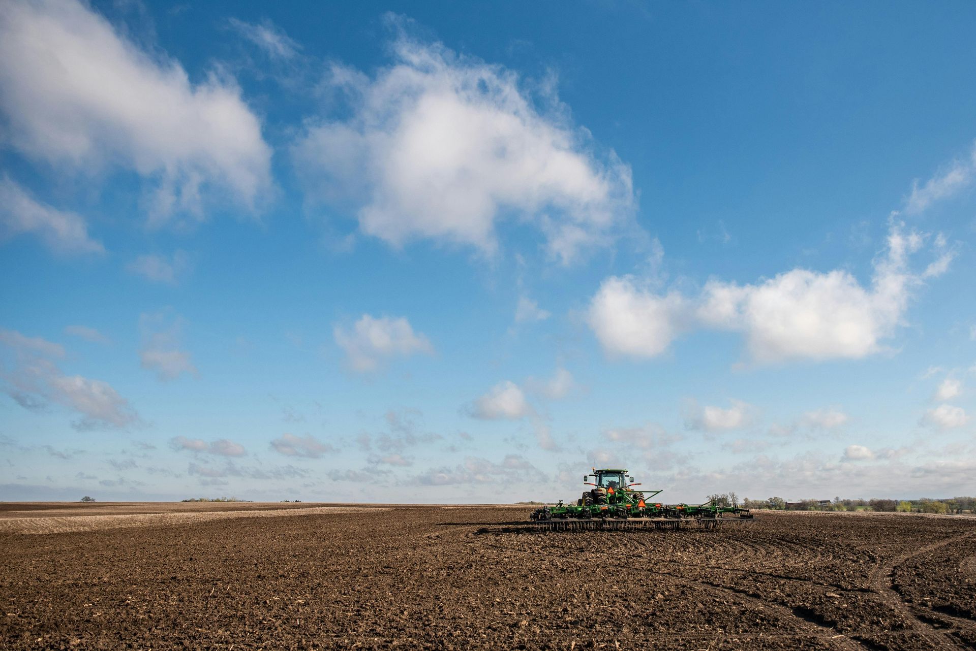 A green tractor pulls a tiller across a vast, dark, freshly plowed field under a bright blue, cloudy sky.