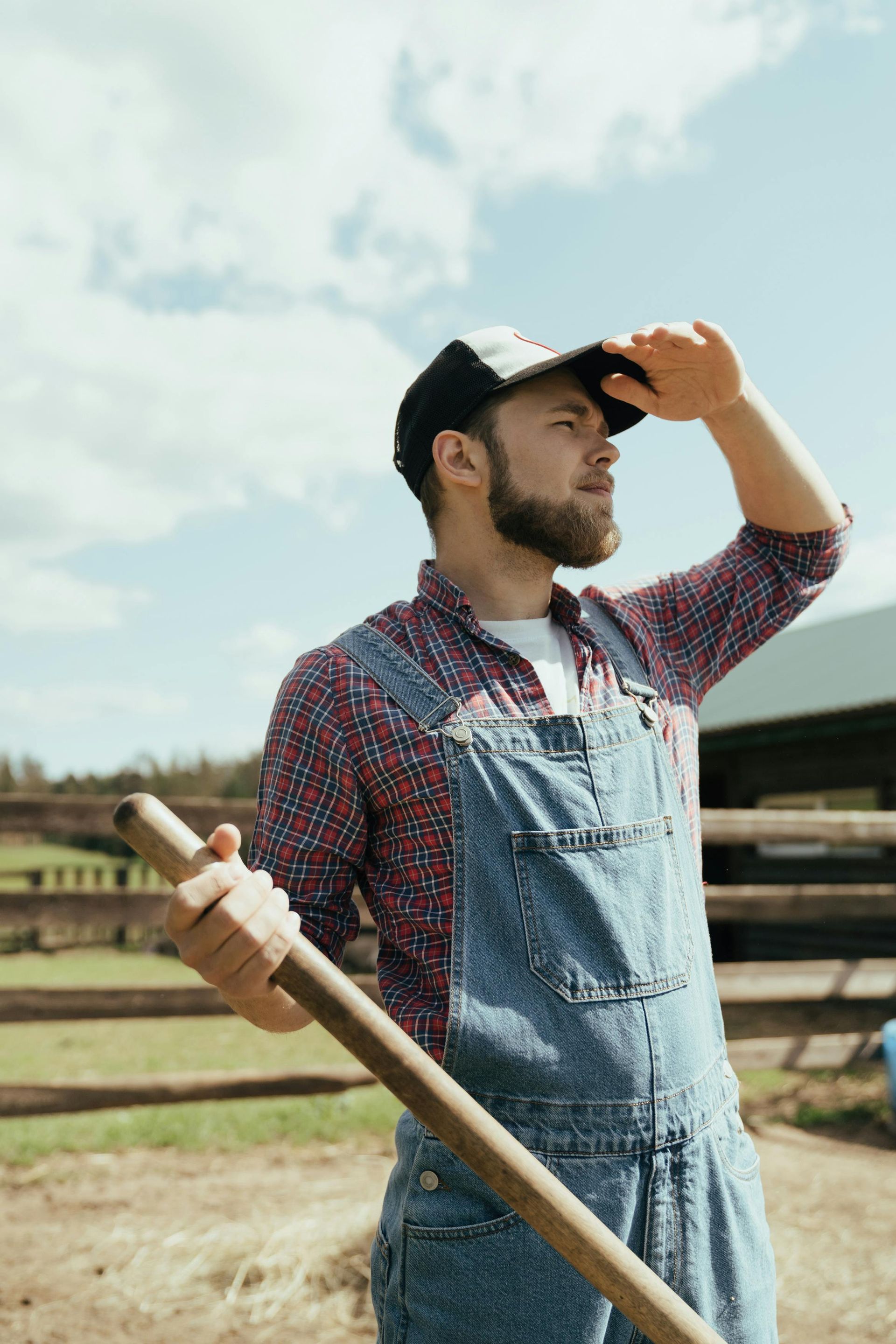A person in overalls and a cap holds a tool and shades their eyes, looking out over a farm on a sunny day.