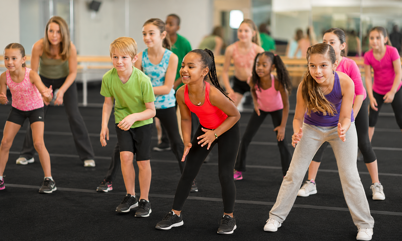 Children doing a group fitness class, leaning forward in a gym with a trainer in the background
