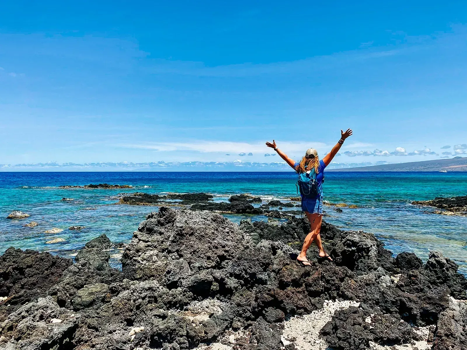 A woman is standing on a rocky beach with her arms in the air.