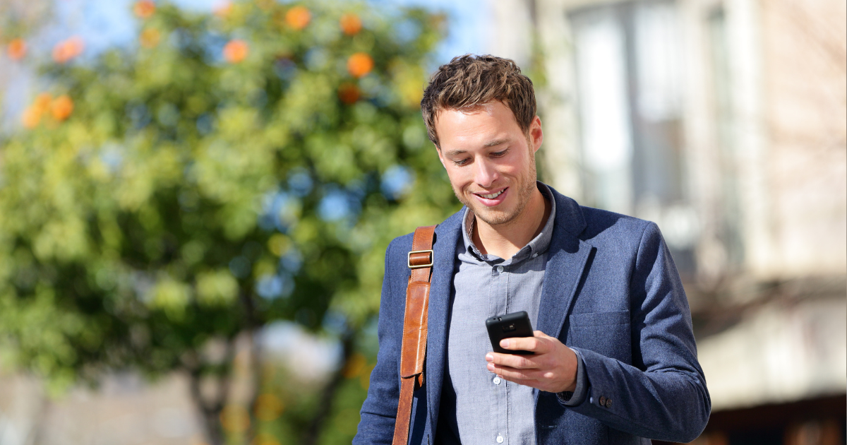 Smiling man in a blue blazer looking at his phone outdoors with a blurred tree and building in the background