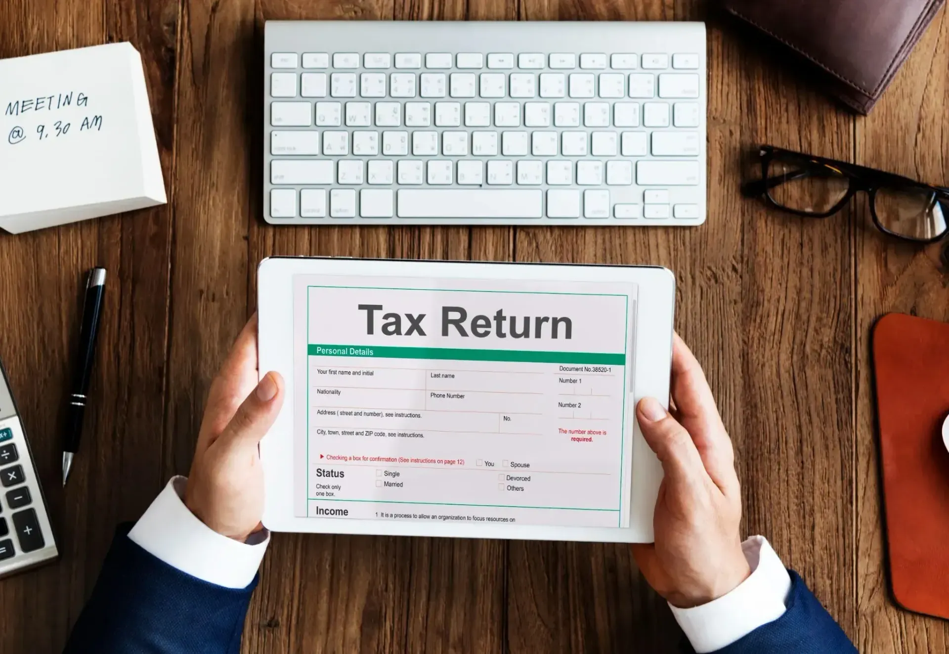Hands holding a tax return form over a wooden desk with a keyboard and calculator nearby