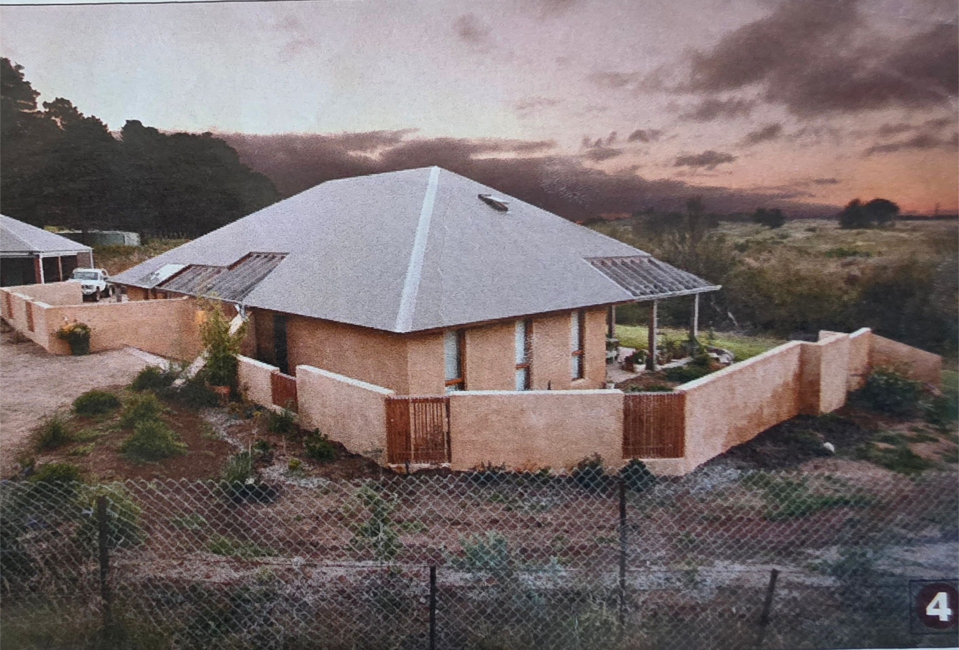 Tan brick house with a flat roof, surrounded by a wall and fence, set in a field.