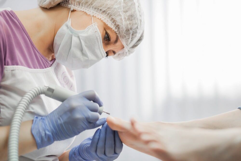 A Woman Wearing A Mask And Gloves Is Polishing A Patient's Feet — Lake Macquarie Podiatry in Belmont, NSW