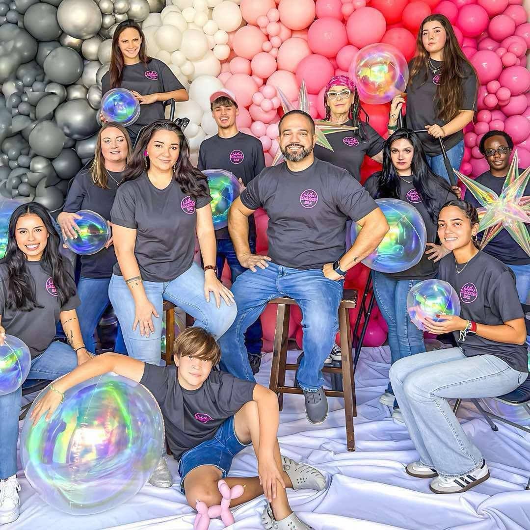 Group of people in grey shirts posing in front of a balloon backdrop, holding iridescent balls.