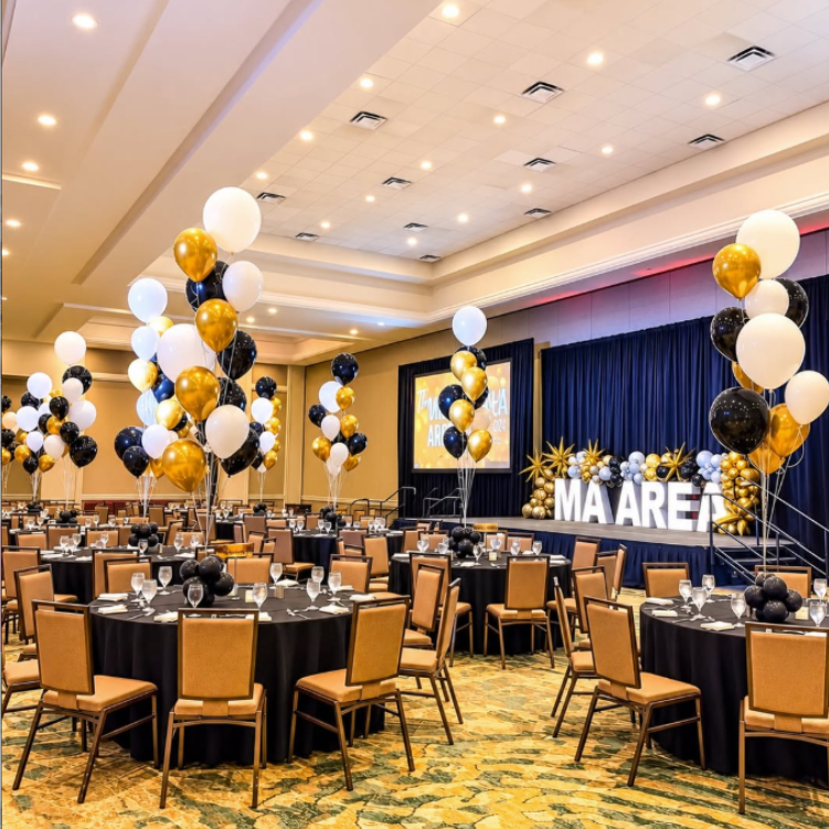 Elegant banquet hall with black, white, and gold balloons, round tables, and a lit stage backdrop