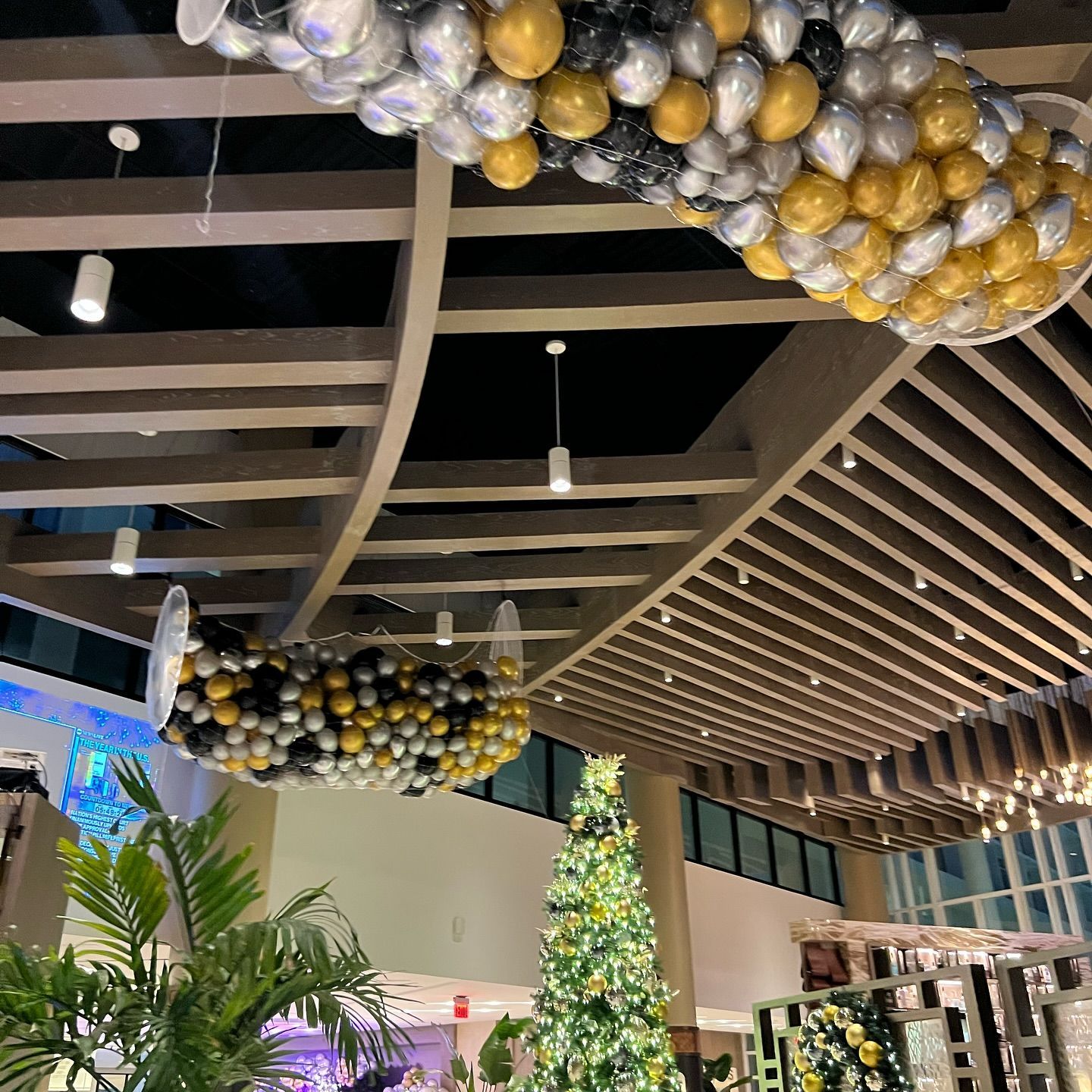 Balloons in gold, silver, and black decorate a building's ceiling. A Christmas tree is visible.
