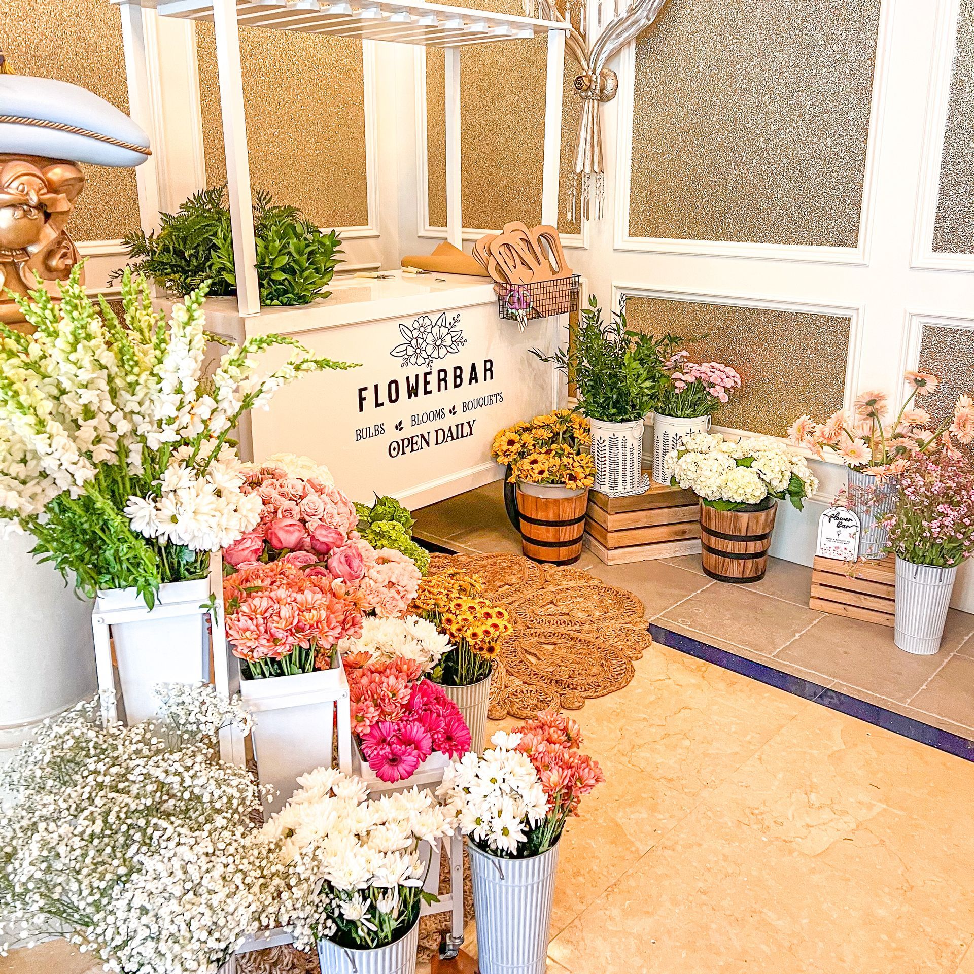 Flower bar display with various flowers, greenery, and decorative elements; white cart and wooden accents.
