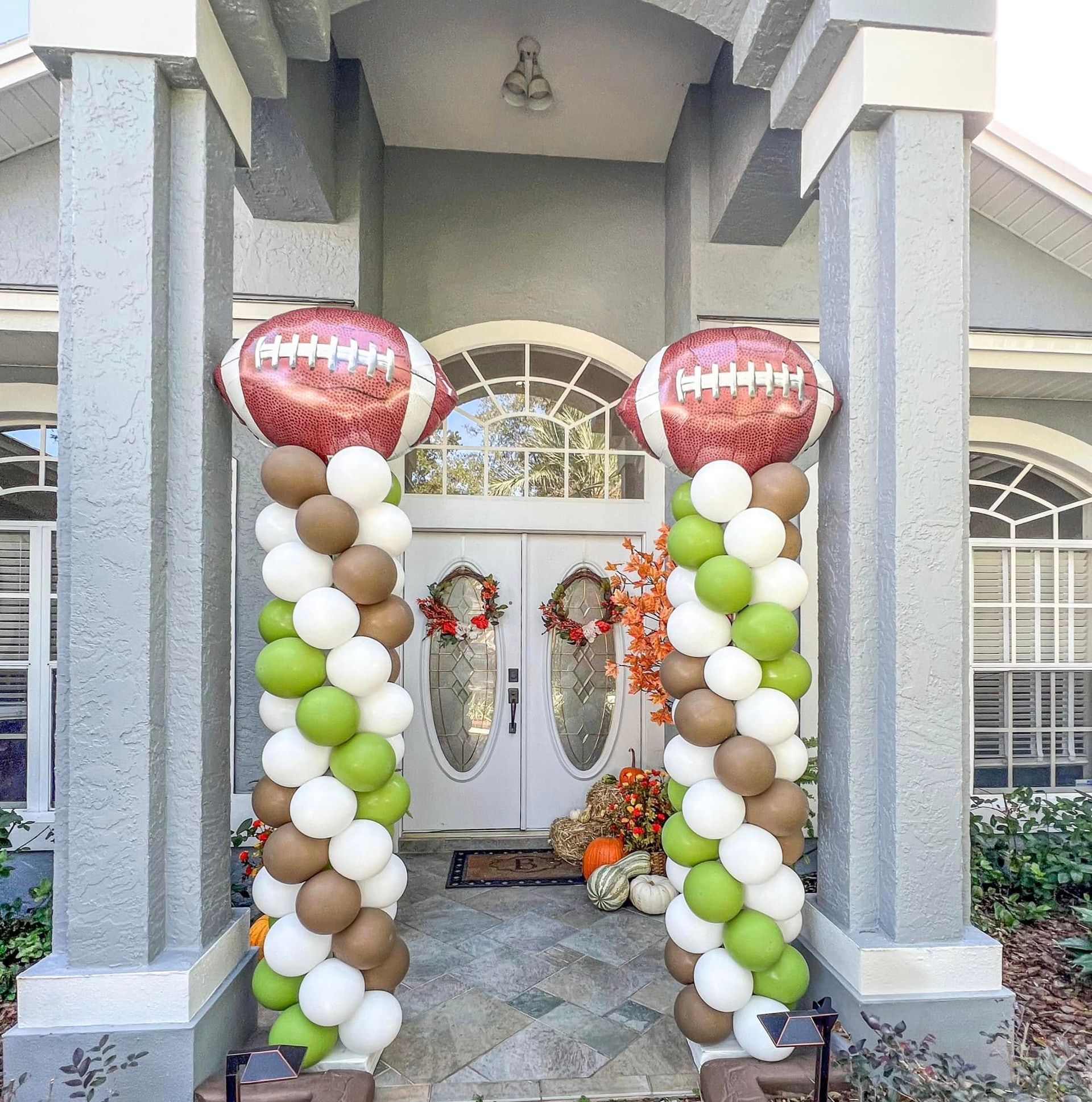 Front porch decorated with white, green, and brown balloon columns and autumn wreaths at double doors.