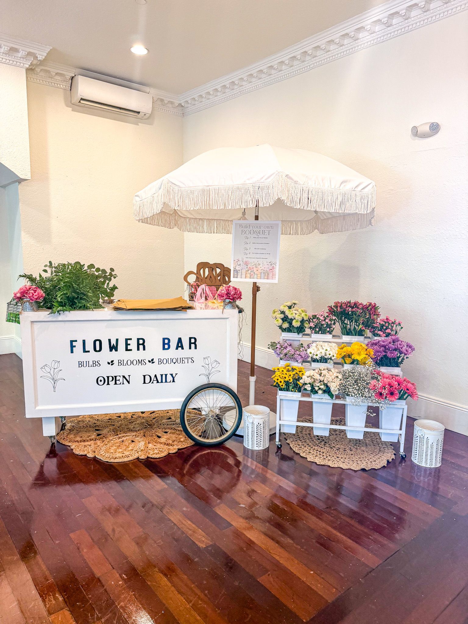 Flower bar with white cart, parasol, and various flowers in a bright room with wood flooring.