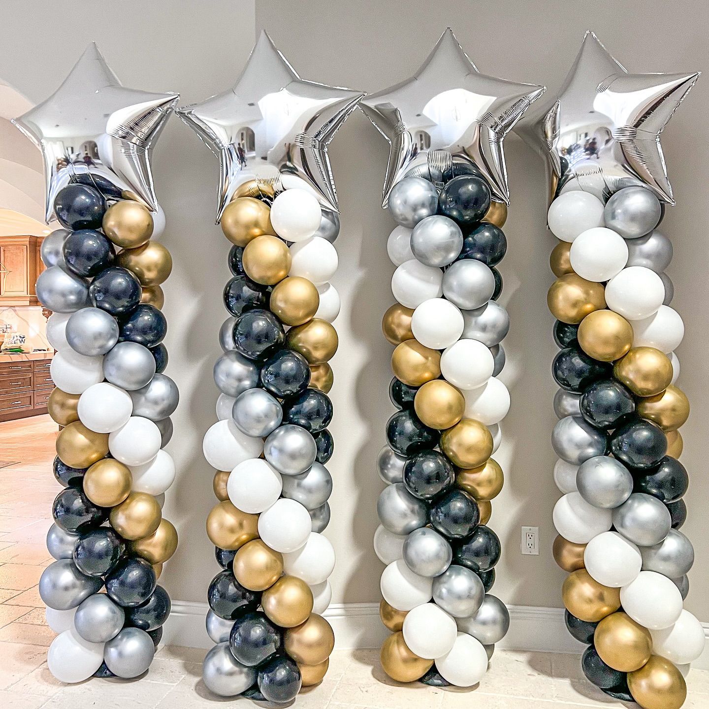 Four balloon columns: silver star toppers, black, silver, gold, and white balloons. Indoor setting.