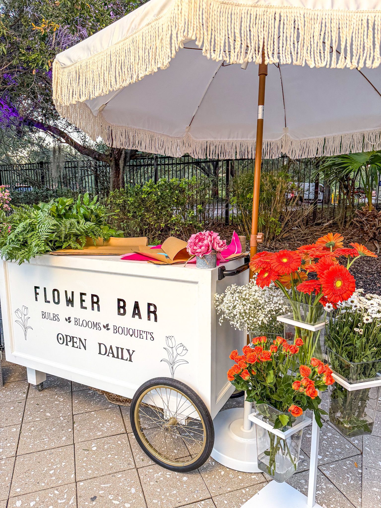 Flower cart under white umbrella, with colorful potted flowers and a sign reading “FLOWER BAR”