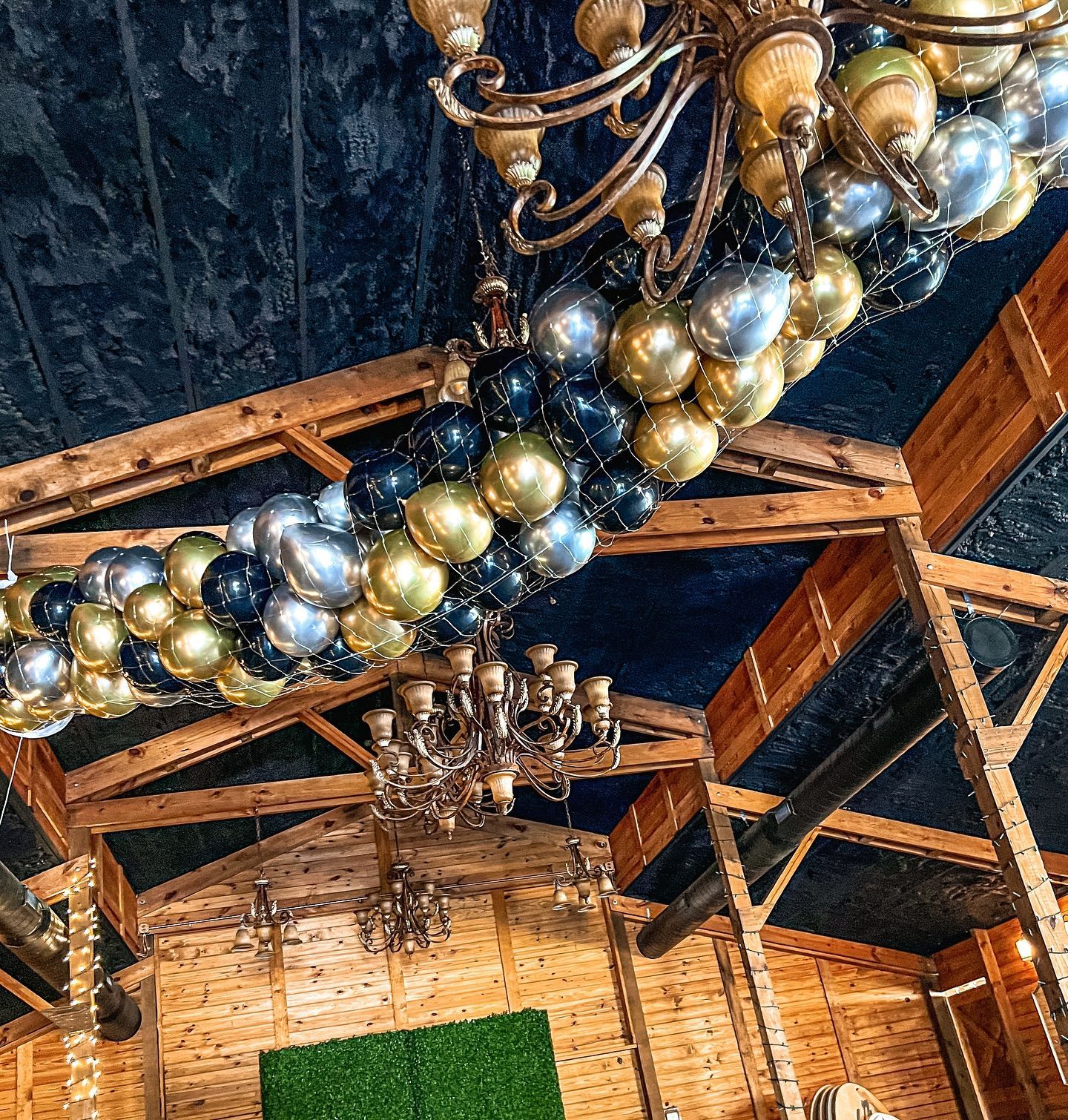 Wooden-beamed interior with blue ceiling, adorned with gold, silver, and black balloons and chandeliers.
