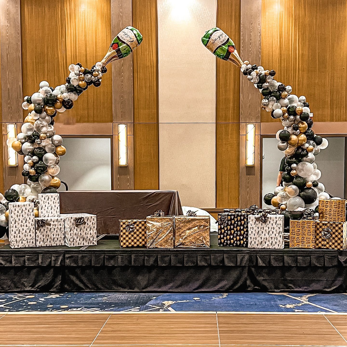Stage setup with gift boxes and champagne bottle balloon arches; black, gold, and silver balloons.