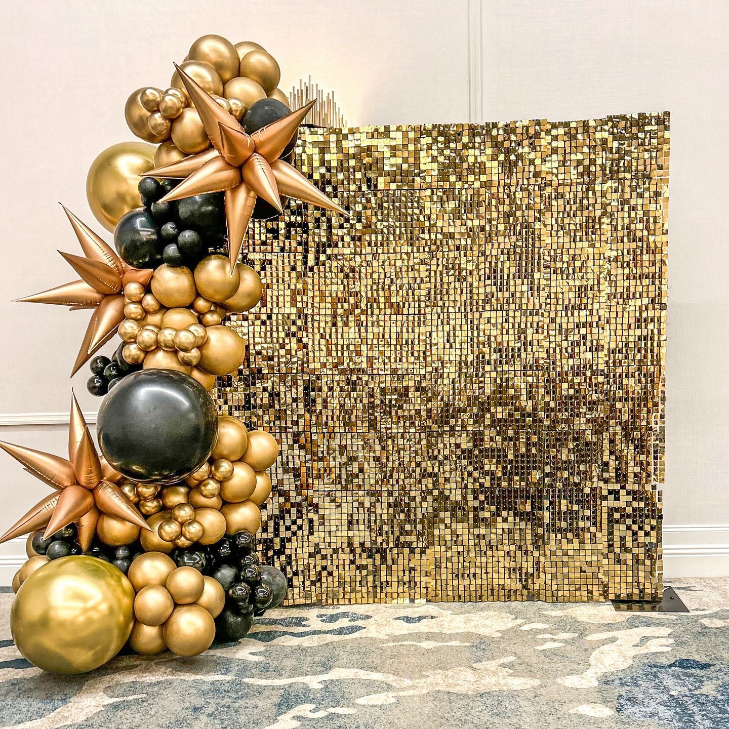 Gold and black balloon arch with star accents next to a golden pixel backdrop on a patterned rug.