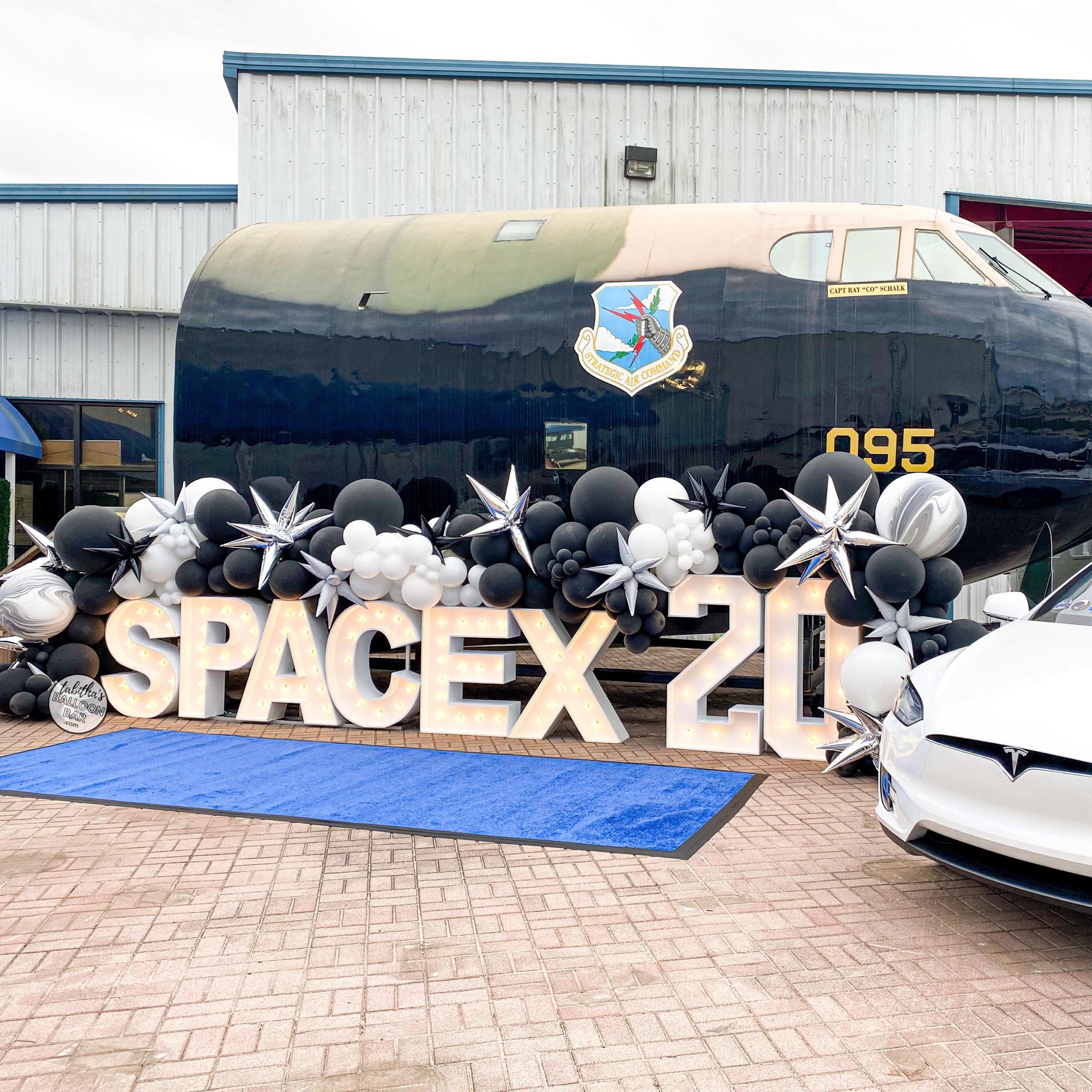 SpaceX 20th anniversary celebration. Black and silver balloons, white Tesla, and airplane in background.
