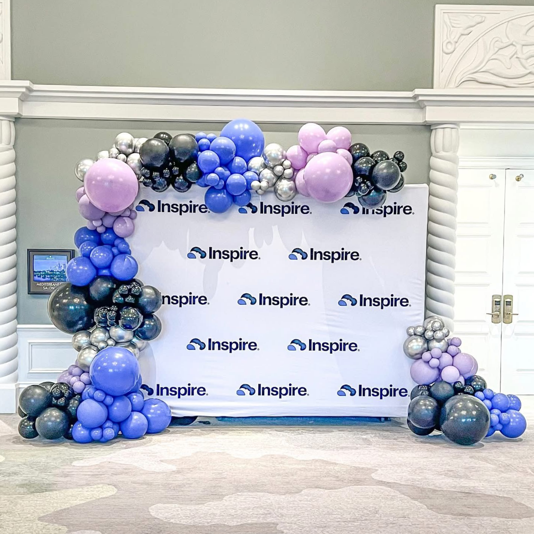 Blue, silver, and lavender balloon arch framing an Inspire logo backdrop at an indoor event entrance