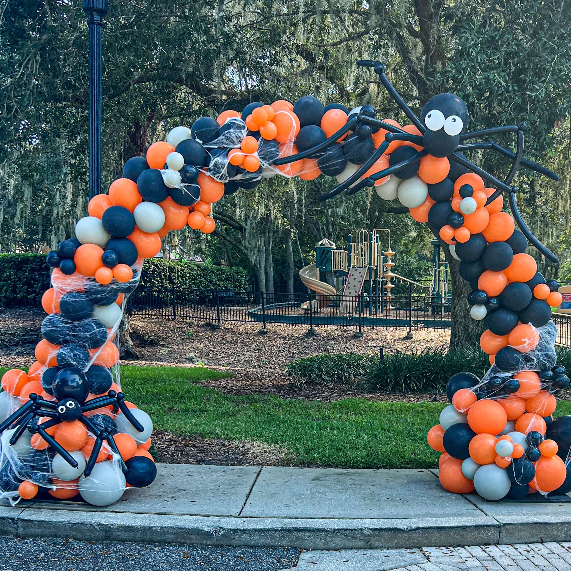 Black and orange balloon arch with spider decorations in a park setting