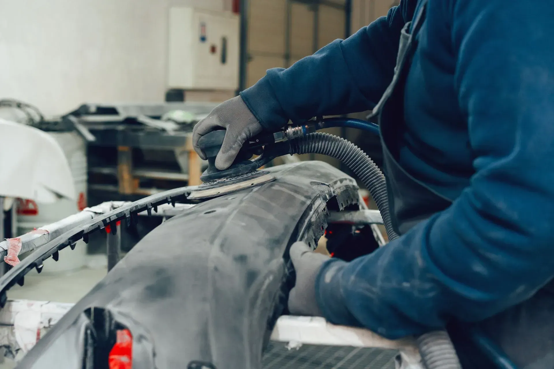 Person in blue sweater using a sander on a car bumper, likely in a workshop.