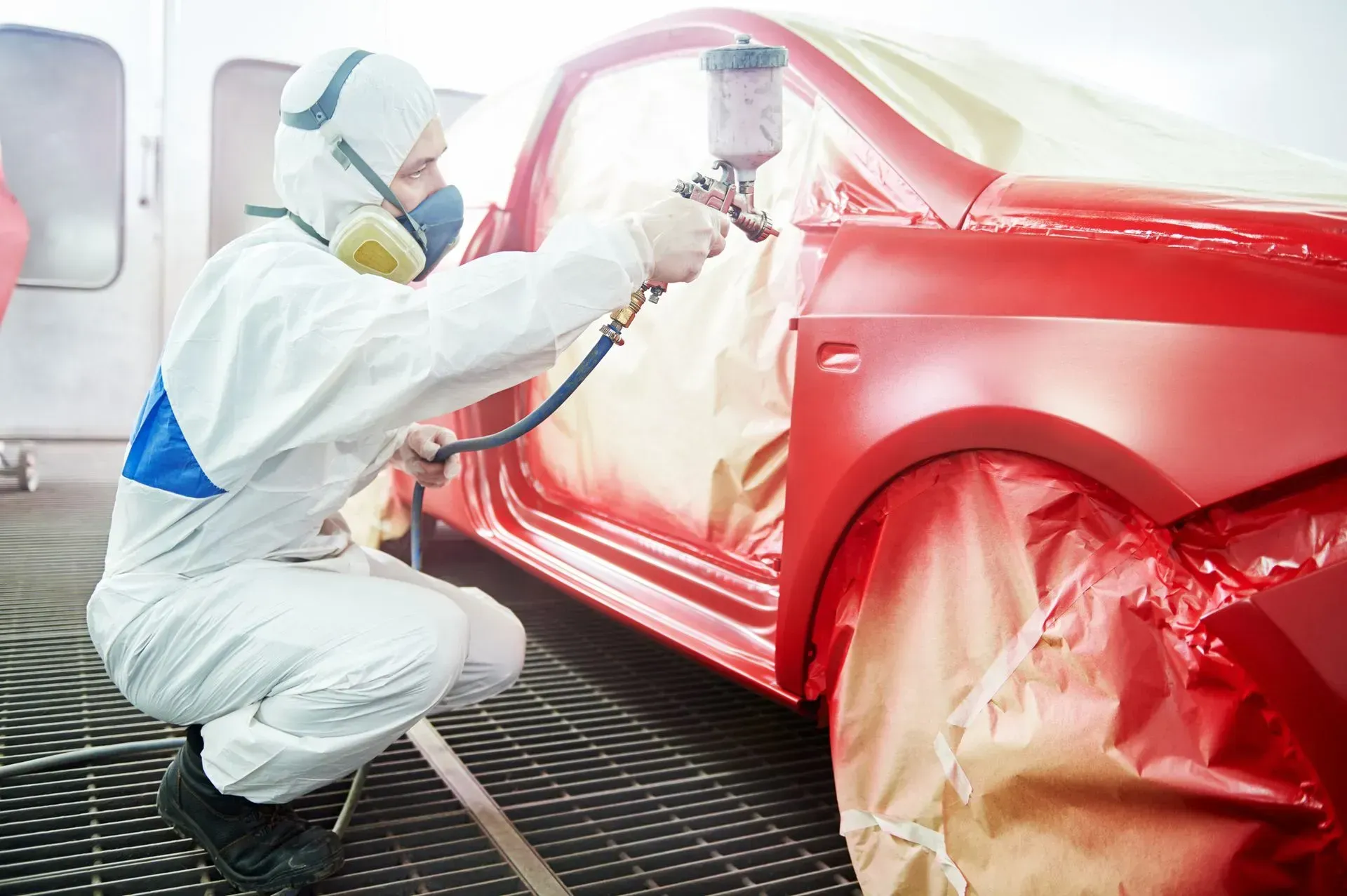 A person in a protective suit paints a red car in a workshop with a spray gun.